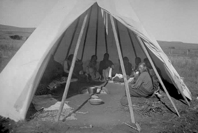 A black and white photo showing 9+ Native Americans gathering in a tepee with a wide open doorway.